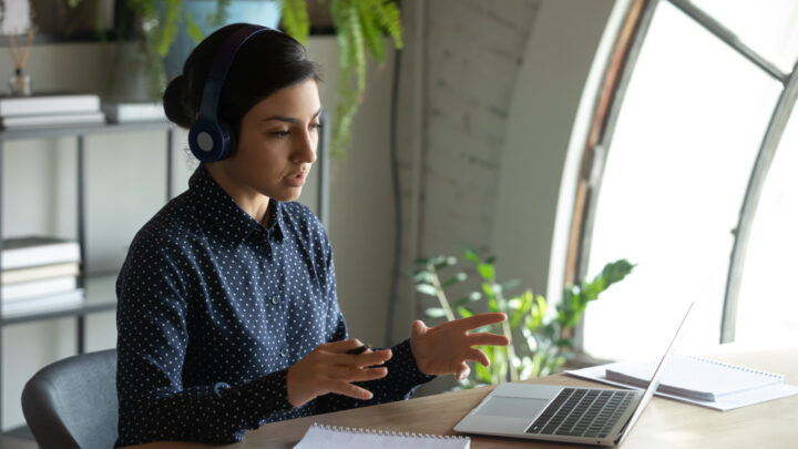 A person wearing headphones sits at a desk with a laptop and notepad, gesturing with one hand, fully immersed in teletherapy.