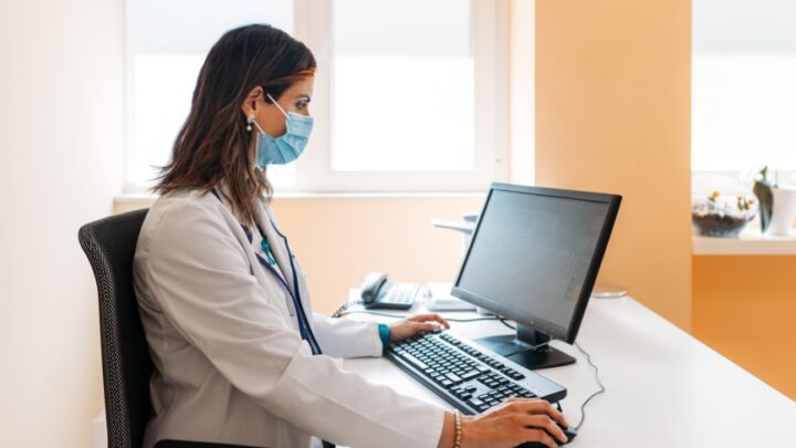 A physician wearing a mask and white coat types on a keyboard at a desk with a computer monitor in the well-lit room, seamlessly blending online communication with medical expertise.