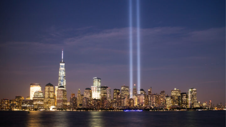 The New York City skyline at night is graced by two vertical beams of light, shining into the sky in remembrance of 9/11, their glow elegantly reflecting in the water.