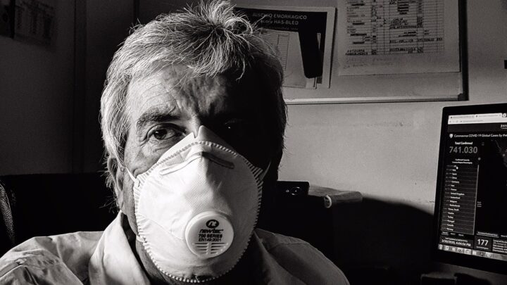 A grayscale image captures a person wearing a white face mask, likely due to the coronavirus, seated in front of a computer screen displaying data, while a bulletin board looms in the background.