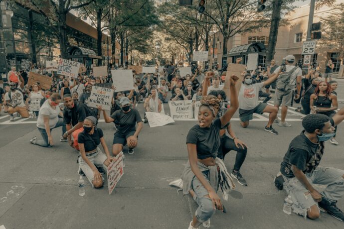 A large group of people kneeling and holding signs in a peaceful protest against racism on a city street, surrounded by trees and buildings.