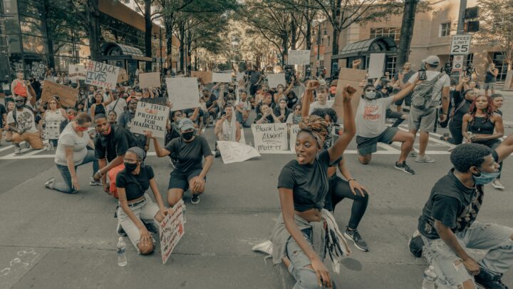 A large group of people kneeling and holding signs in a peaceful protest against racism on a city street, surrounded by trees and buildings.