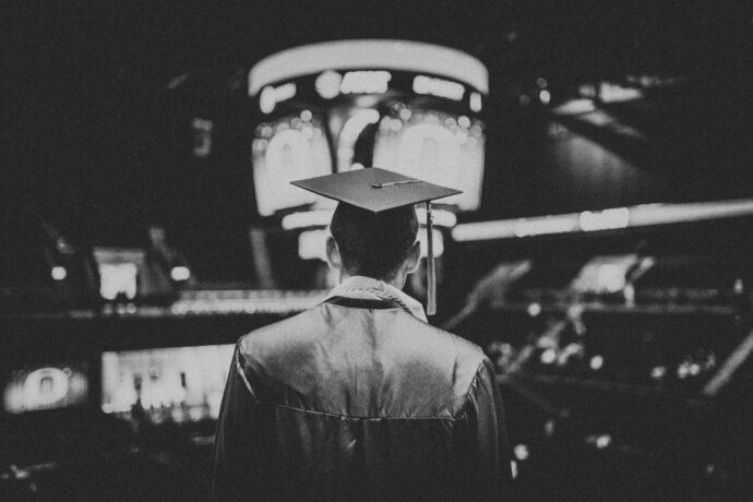 A med student in a cap and gown stands facing a large screen in a dimly lit arena, reflecting on graduating early during the COVID-19 pandemic.