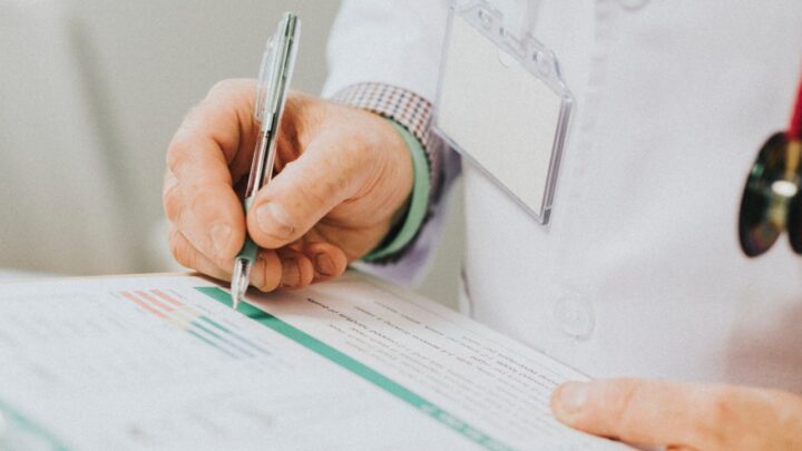 A person in a white coat holds a pen, meticulously updating medical records on a clipboard.