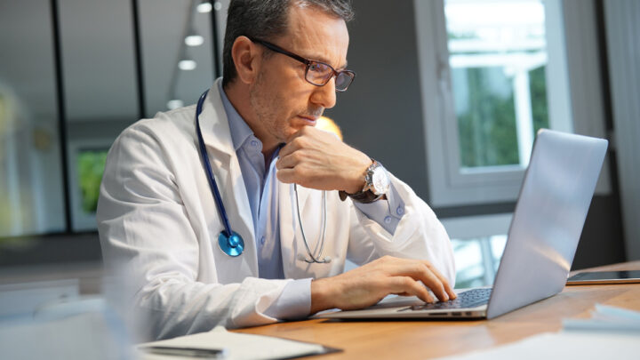 A dedicated doctor in a white coat and stethoscope sits at a desk, concentrating on a laptop in the well-lit office, providing essential support to fellow healthcare workers navigating the challenges of COVID.