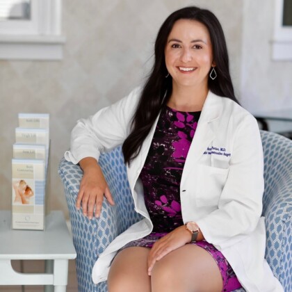 Dr. Anna Pavlov, MD, sits on a patterned chair in a white medical coat, smiling warmly at the camera. Pamphlets are displayed on a stand next to her.