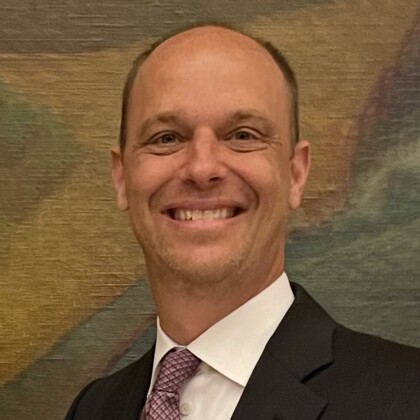 Dr. Travis Hiles, MD, smiles at the camera in a suit and tie, standing in front of a blurred, textured background.