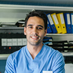 A man in blue medical scrubs stands in front of shelves with organized binders and files, smiling at the camera, ready to share advice on websites for doctors.
