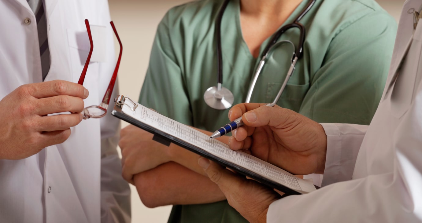 Three healthcare professionals are in discussion, with one holding a pen and clipboard while another clutches eyeglasses. A stethoscope drapes around one individual's neck as they focus on end-of-life care strategies.