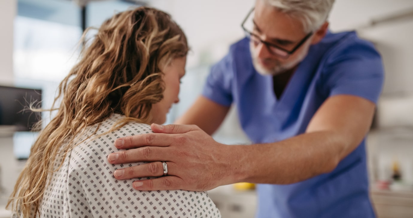 A medical professional in blue scrubs places a comforting hand on the shoulder of a patient wearing a hospital gown, embodying compassionate cancer care within an examination room.