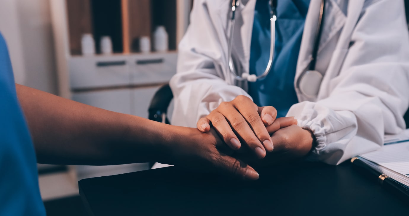 A doctor in a white coat holds a patient's hand reassuringly over the desk, surrounded by medical supplies. The atmosphere speaks to the ethical considerations inherent in cancer care, where compassion and understanding are as vital as any treatment.