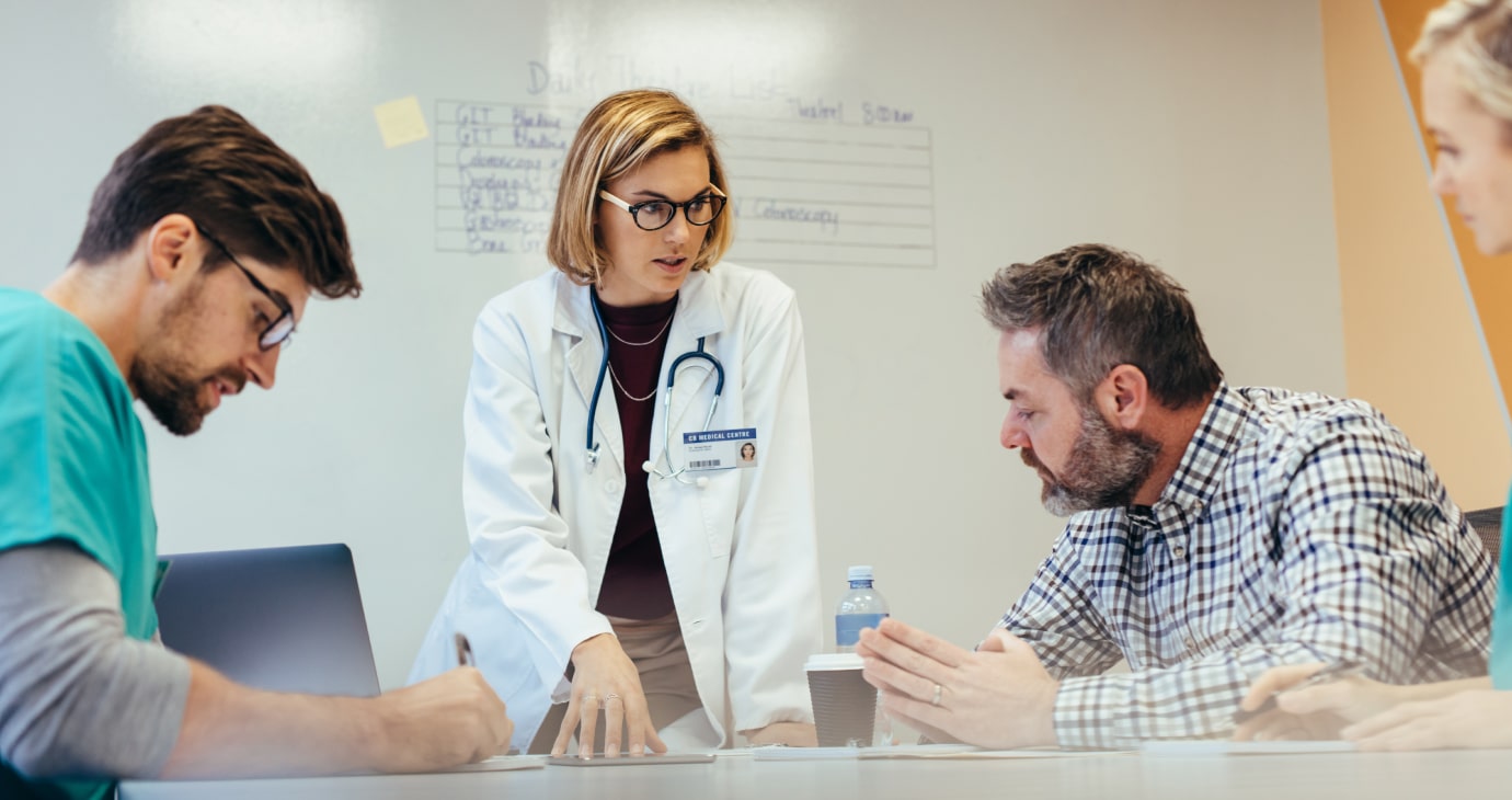 A doctor in a lab coat discusses ethical considerations with three colleagues seated at a table, focused intently on a document in the meeting room.