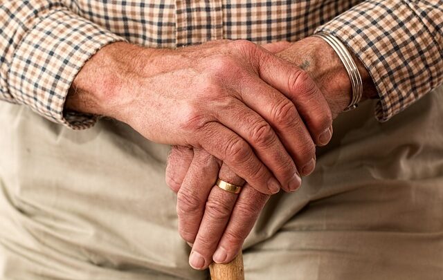 Close-up of an elderly person's hands resting on a sturdy wooden cane, wearing a checkered shirt and a gold ring—essentials for supporting posture and maintaining back health.
