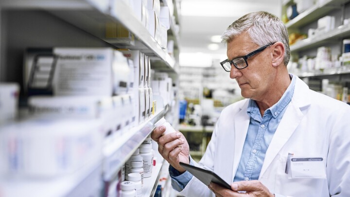 A pharmacist wearing a white lab coat and glasses examines a medicine bottle while holding a tablet in a pharmacy aisle, reflecting pharmacist trends and the role expansion within the profession.