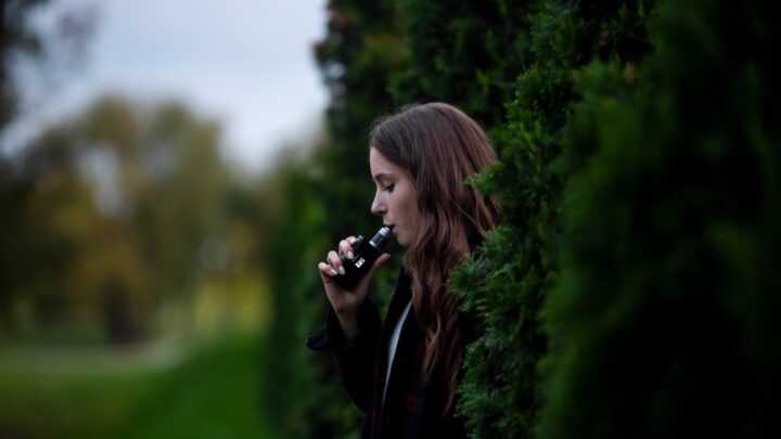 A person with long hair is outdoors, standing near tall greenery, holding and using e-cigarettes with curiosity.