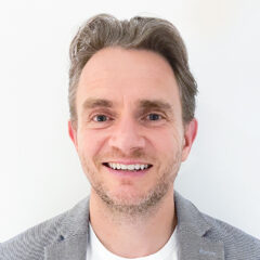 Smiling person with short, styled hair, wearing a grey blazer and white shirt, against a plain white background.