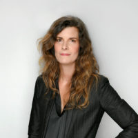Erin Fitzgerald, a woman with long, wavy hair, elegantly wears a black blazer as she stands against a plain background.
