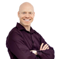 Guy Jones, sporting a shaved head and wearing a dark long-sleeve shirt, stands confidently with arms crossed, smiling warmly at the camera against a plain white background.