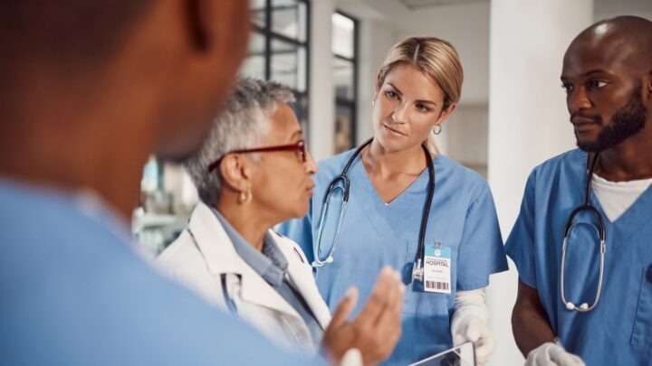 Medical professionals in scrubs have a discussion in a hospital setting. One person gestures while others, including experienced physicians and NPs, listen attentively, wearing stethoscopes and ID badges. The focus shifts to the ongoing "ill-trained NPs" controversy affecting team dynamics.