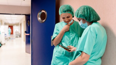 Two healthcare professionals in scrubs and hairnets review information on a smartphone in a hospital corridor, sharing insights from physician experiences.
