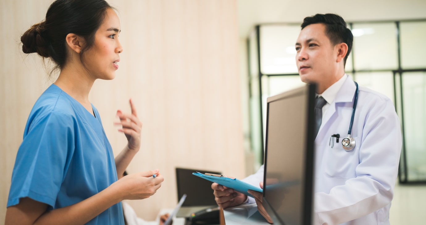 A healthcare professional in blue scrubs talks to a physician in a white coat holding a clipboard, focusing on how to improve patient satisfaction in a medical office setting.