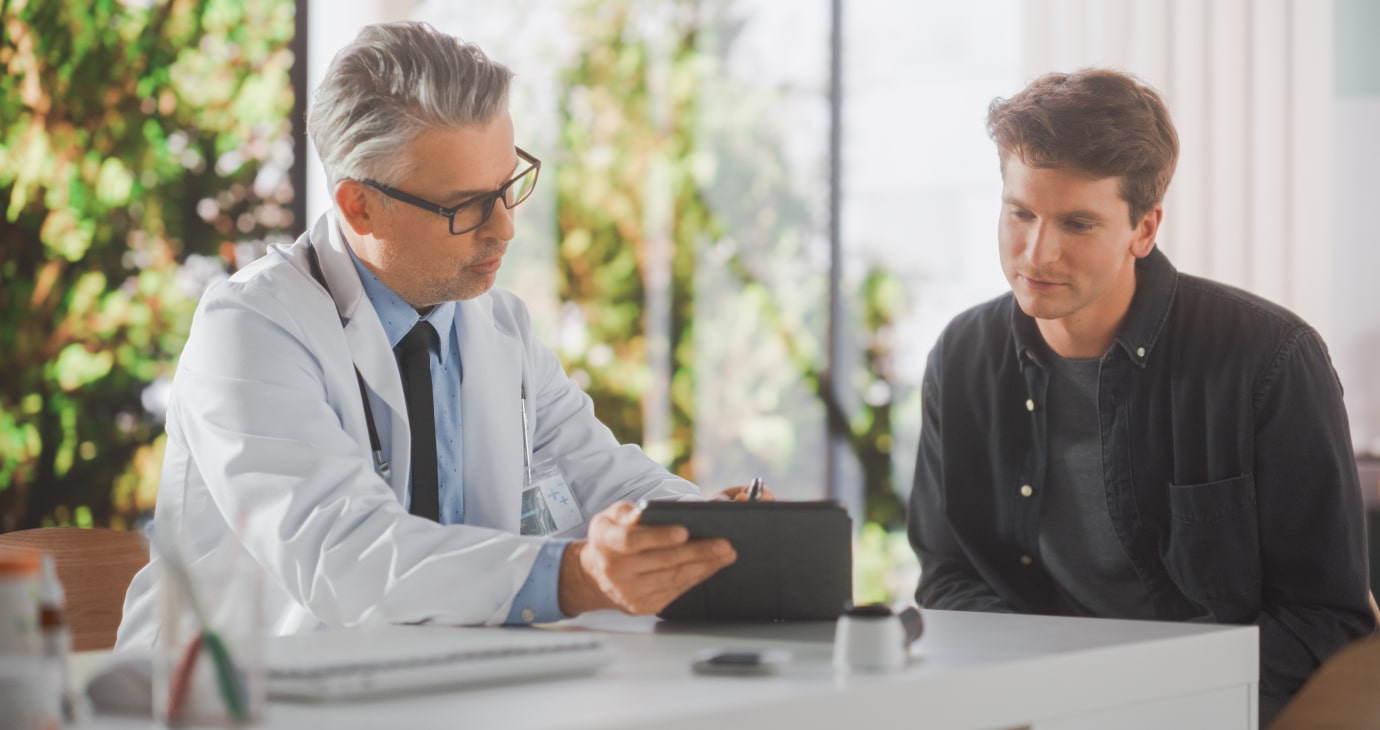 A doctor and a patient sit at a desk, reviewing information on a tablet in a bright office with large windows and plants outside. Their collaborative approach aims to improve patient satisfaction, fostering trust and understanding.