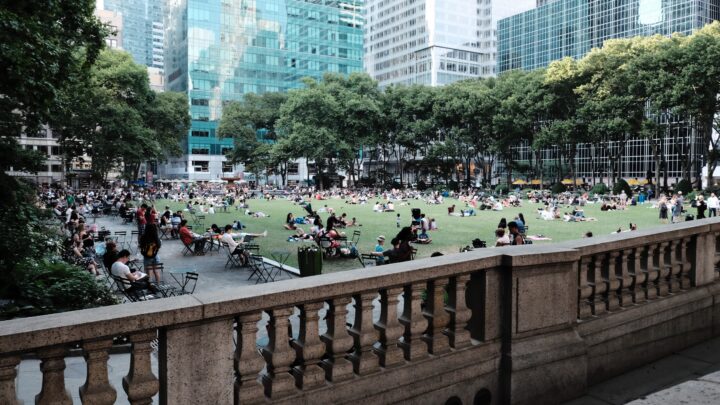 People relaxing on the grass and sitting on chairs in a city park, surrounded by tall buildings and trees, enjoy conversations about Sermo while appreciating the collective effort towards herd immunity.