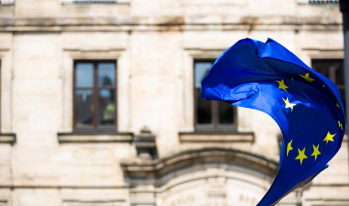 The EU flag gracefully waves in front of a historic building with arched windows, symbolizing unity even as the region faces challenges like high infections.