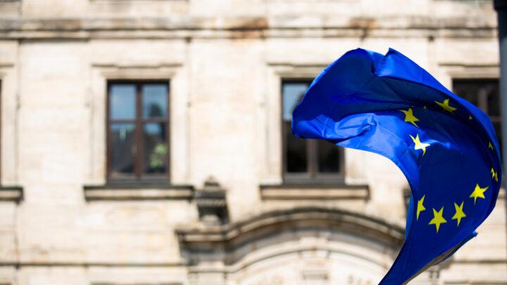 The EU flag gracefully waves in front of a historic building with arched windows, symbolizing unity even as the region faces challenges like high infections.