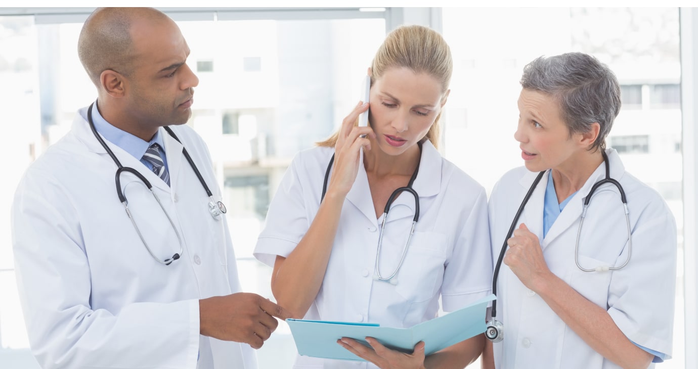 Three healthcare professionals in white coats, two women and one man, confer over a clipboard. One woman is on a phone call. Stethoscopes around their necks signify their dedicated leadership in navigating medical challenges.