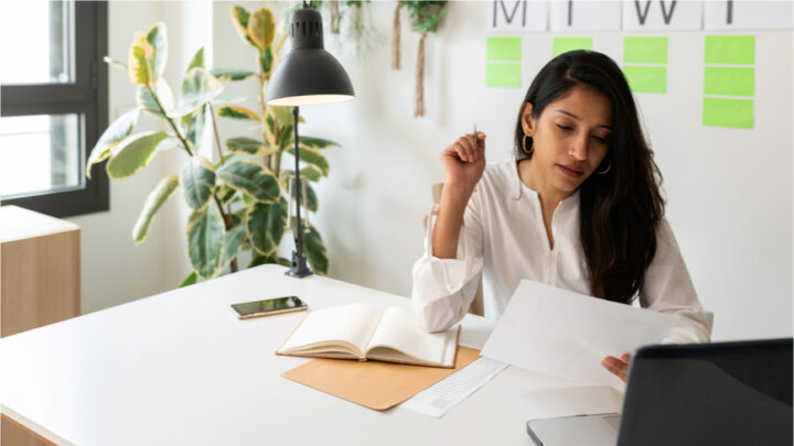 A woman in a white shirt sits at her desk, reviewing a document on copay programs. A laptop, notebook, and phone are on the desk, while a lamp and plant add warmth to the workspace.