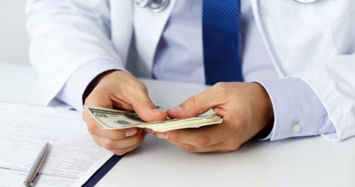 A doctor in a white coat reviews physician compensation while counting a stack of cash over a desk cluttered with paperwork and a pen.