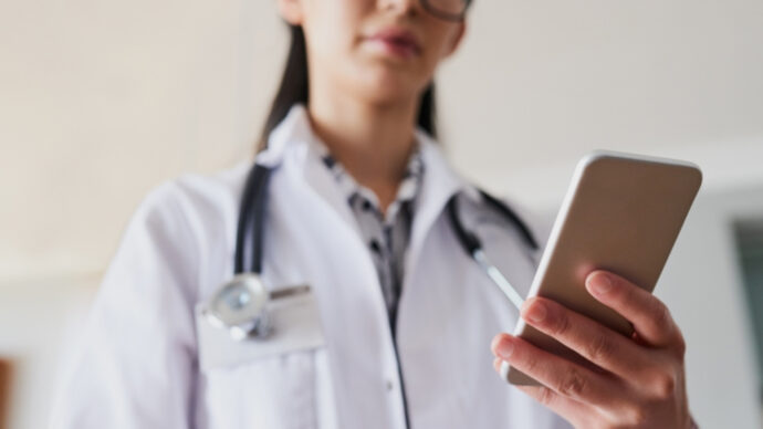 A person in a white coat with a stethoscope around their neck is holding a smartphone, possibly checking the latest updates on the 2024 presidential election.