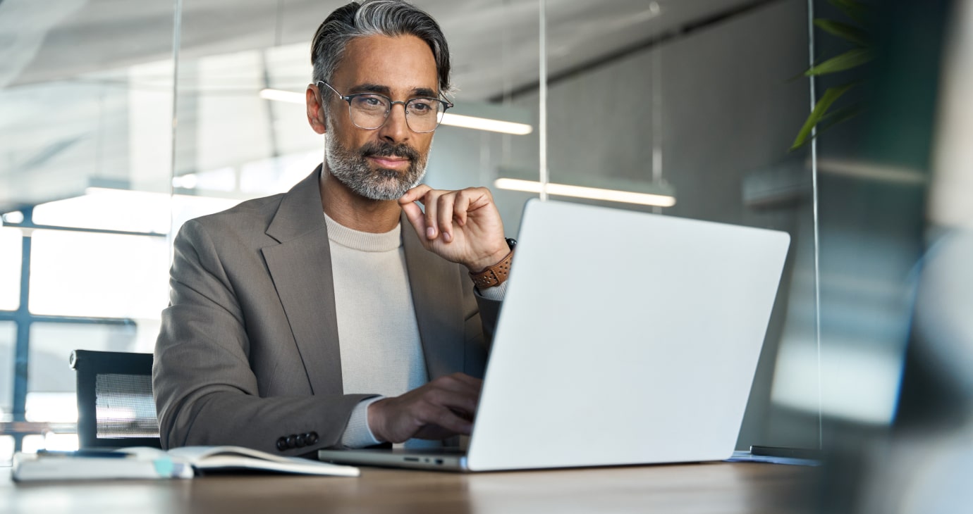 A seasoned physician with gray hair and glasses works on a laptop in a sleek, modern office. A notebook and pen lie on the table beside him, perhaps holding notes on his latest venture into real estate investing.