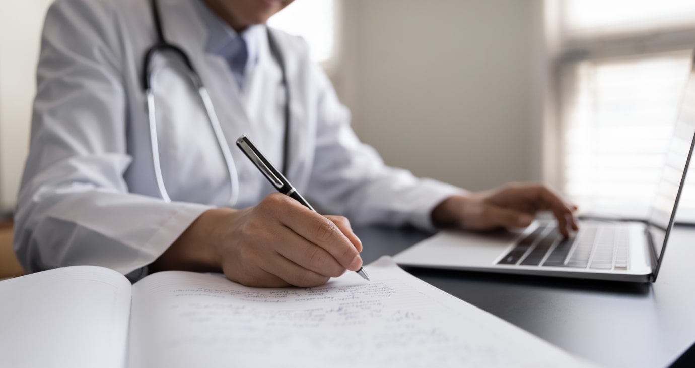 A physician in a white coat writes on paper while using a laptop, with a stethoscope around their neck, carefully balancing patient care with real estate investing strategies.