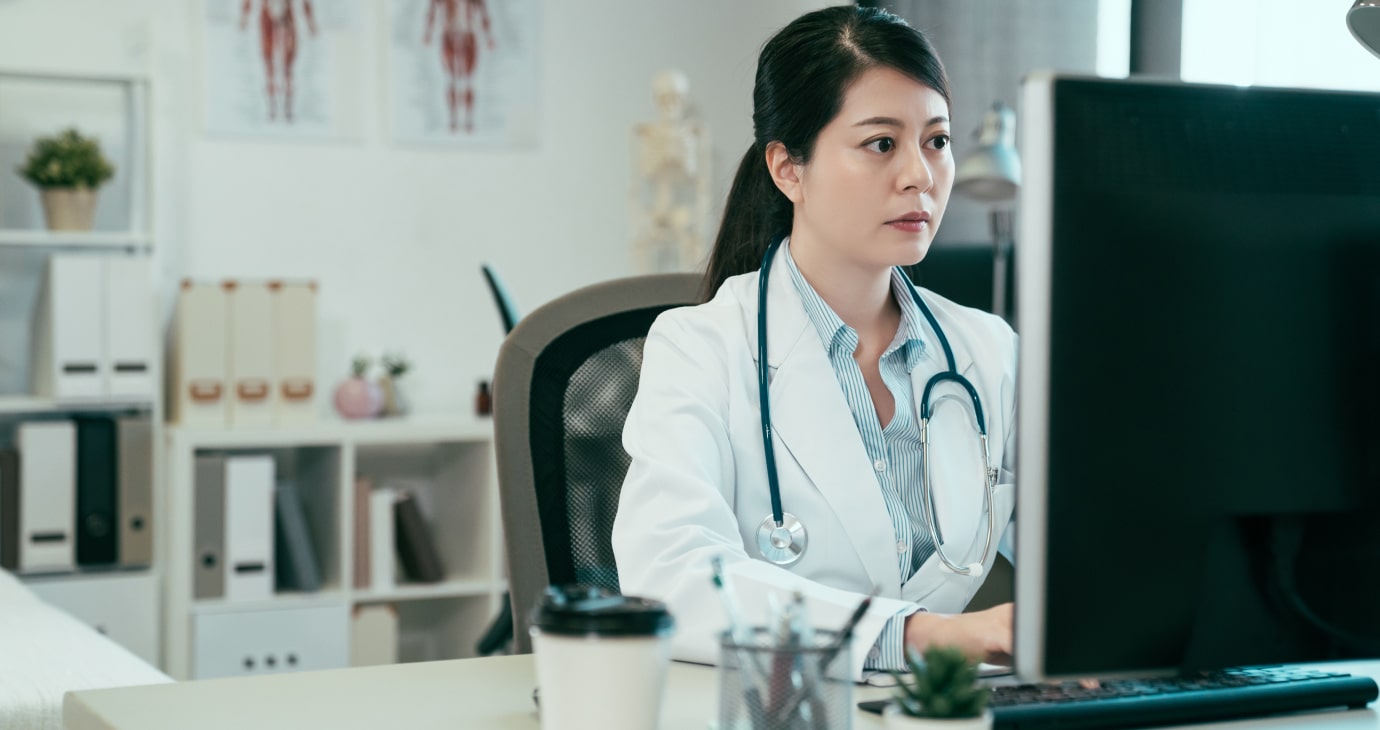 A physician in a white coat is working on a computer at a desk, surrounded by a coffee cup, bookshelf, and anatomical posters, while contemplating the potential of real estate investing.