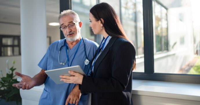 A doctor in blue scrubs chats with a woman in a suit, who is holding a tablet and discussing real estate investing, as they stand by the window.