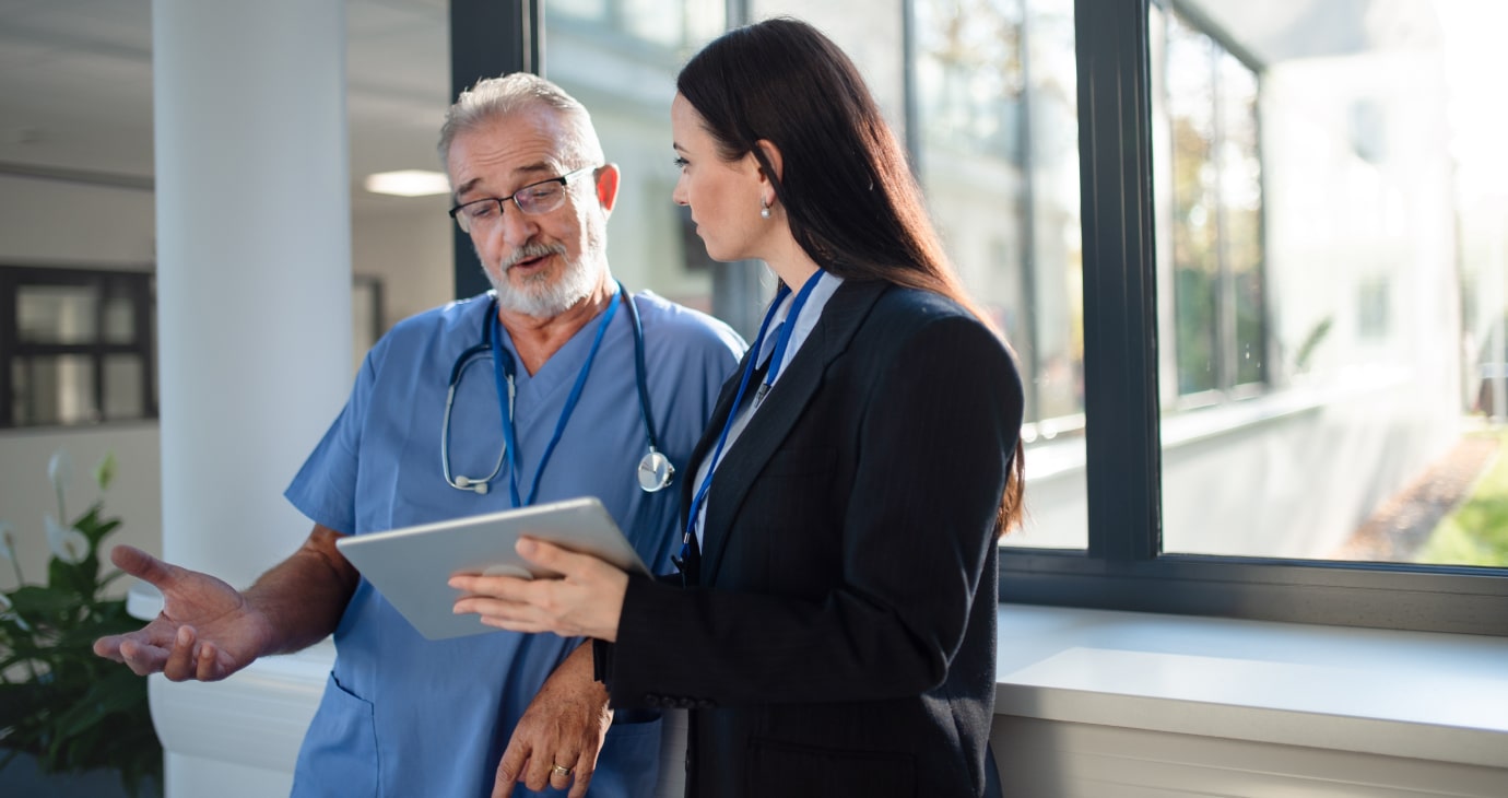 A physician in blue scrubs chats with a woman in a suit, who is holding a tablet and discussing real estate investing, as they stand by the window.