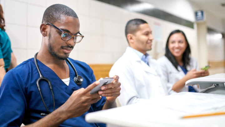 A healthcare professional in navy scrubs sits and uses a smartphone, possibly exploring social media impact, as two colleagues in white coats smile and converse in the background.