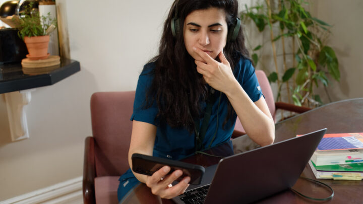 A woman wearing headphones sits at a table with a laptop, holding a phone and looking contemplative. Books are stacked on the table beside her as she explores data-driven insights.