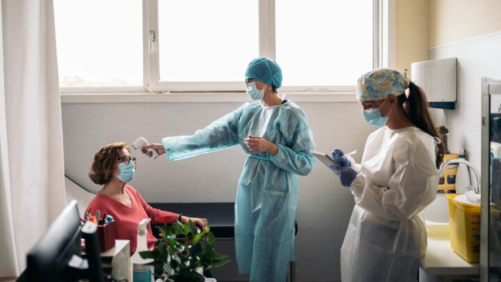 Healthcare workers in protective gear check a seated woman's temperature in a medical room, signaling a cautious return to pre-pandemic patient activity.