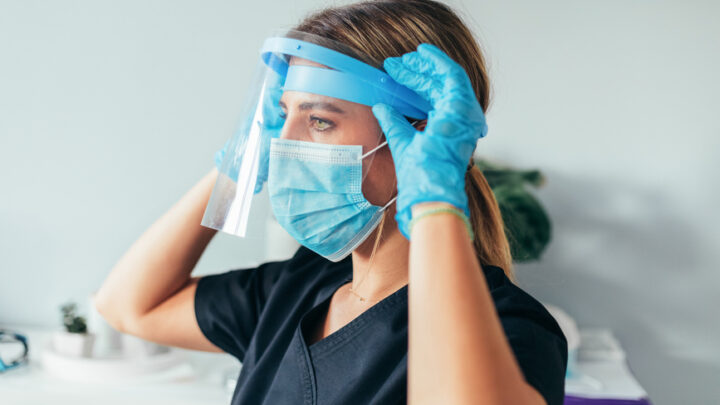 A healthcare worker wearing a face mask and blue gloves adjusts a face shield, exemplifying the shift towards telehealth adoption.