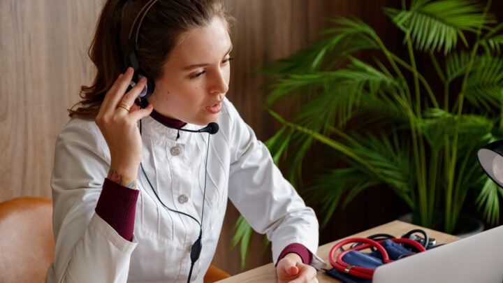 A person in a white coat uses a headset while looking at a laptop, likely engaging in remote engagement. A stethoscope sits on the table beside them, with a potted plant adding a touch of nature to the background.