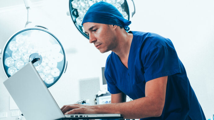 In a modern operating room illuminated by circular lights, a person in blue surgical attire is intently focused on using a laptop, harnessing AI technology to enhance precision and decision-making during procedures.