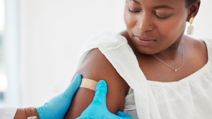 A person in a white top looks at a band-aid on their upper arm as a healthcare provider in blue gloves attends to it after administering the shingles vaccine.