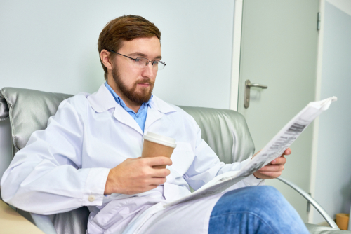 A person in a lab coat sits on a couch, sipping coffee while engrossed in the newspaper headlines.
