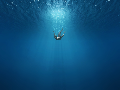 A person floats serenely underwater, surrounded by the deep blue ocean with sunlight filtering through, creating an atmosphere so tranquil that it feels as if they are momentarily drowning in peace.