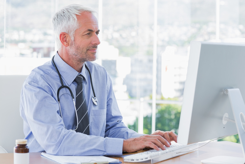 A male doctor with gray hair and a stethoscope around his neck types on a computer in his bright office, managing his online reputation. A bottle and notepad rest on the desk beside him.