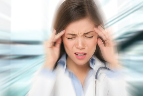 A woman in a doctor's coat is clutching her head with both hands, eyes closed, clearly experiencing pain or stress. The background is blurred.
