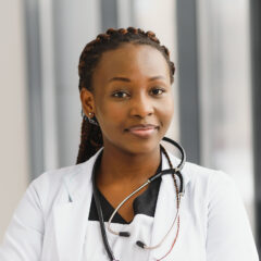 A person in a white coat with a stethoscope around their neck stands indoors, reflecting on the potential of a side hustle for doctors while looking at the camera.